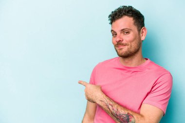 Mavi arka planda izole edilmiş genç beyaz adam gülümsüyor ve boş alanda bir şeyler gösteriyor..