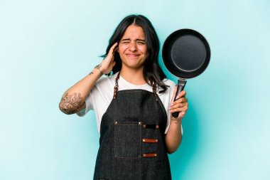 Young hispanic cooker woman holding frying pan isolated on blue background covering ears with hands.