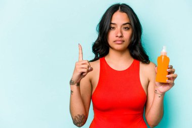 Mavi arka planda güneş kremi tutan genç İspanyol kadın parmağıyla bir numarayı gösteriyor..