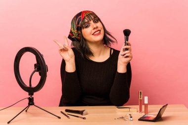 Makyaj eğitmeni genç bir kadın pembe arka planda izole edilmiş neşeli ve kaygısız bir barış sembolünü parmaklarıyla gösteriyor..