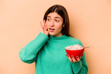 Latin kökenli genç bir kadın bej arka planda izole edilmiş mısır gevreği yiyor ve bir dedikoduyu dinlemeye çalışıyor..