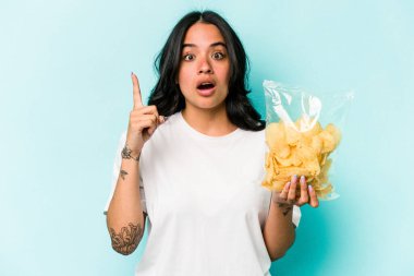 Mavi arka planda izole edilmiş bir paket cipsi tutan genç İspanyol kadının bir fikri, ilham verici bir fikri var..