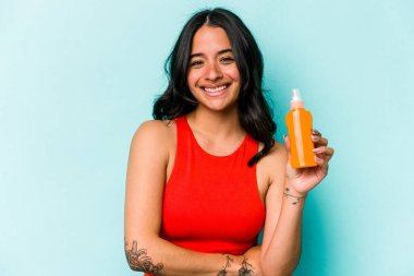 Mavi arka planda güneş kremi tutan genç İspanyol kadın gülüyor ve eğleniyor..