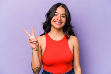 Mor arka planda izole edilmiş genç İspanyol kadın zafer işareti gösteriyor ve gülümsüyor..