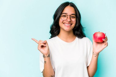 Mavi arka planda izole edilmiş bir elma tutan genç İspanyol kadın gülümsüyor ve boş alanda bir şey gösteriyor..