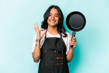 Young hispanic cooker woman holding frying pan isolated on blue background cheerful and confident showing ok gesture.