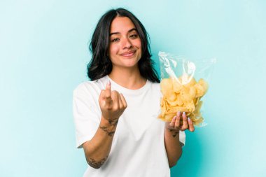 Genç İspanyol kadın mavi arka planda izole edilmiş bir çanta tutuyor. Sanki seni davet ediyormuş gibi parmağıyla işaret ediyor..