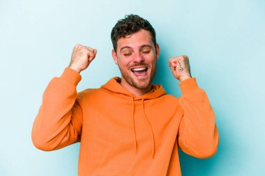 Mavi arka planda izole edilmiş genç beyaz bir adam zaferi, tutkuyu ve coşkuyu kutluyor, mutlu bir ifade..