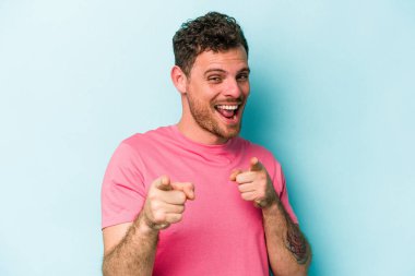 Mavi arka planda izole edilmiş genç beyaz adam parmaklarıyla ön tarafı işaret ediyor..