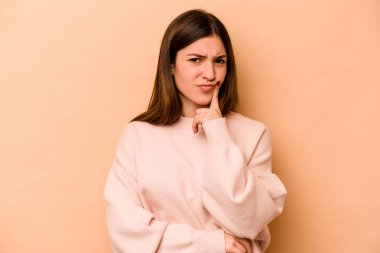 Latin kökenli genç bir kadın bej arka planda izole edilmiş. Mutsuz ve alaycı bir ifadeyle kameraya bakıyor..