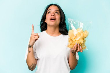 Genç İspanyol kadın elinde bir çanta dolusu cipsle mavi arka planda tek başına ağzı açık bir şekilde üst tarafı işaret ediyor..