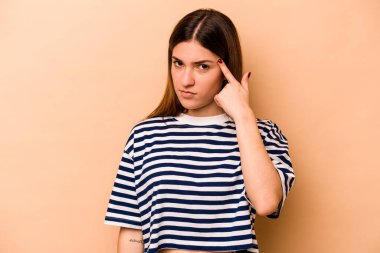 Latin kökenli genç bir kadın, bej arka planda izole edilmiş, parmağıyla tapınağı işaret ederek, düşünerek, bir göreve odaklanmış..