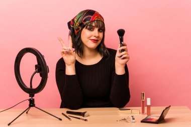 Genç makyajcı kadın makyaj dersi veriyor. Pembe arka planda iki numarayı parmaklarıyla gösteriyor..