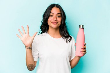 Mavi arka planda termosu olan genç İspanyol kadın gülümseyerek beş numarayı parmaklarıyla gösteriyor..