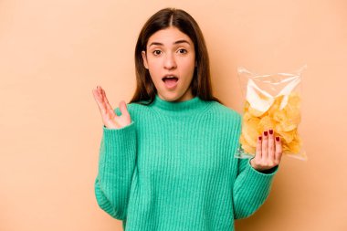 Latin kökenli genç bir kadın elinde bir paket cipsle bej arka planda mahsur kaldı. Şaşırdı ve şok oldu..