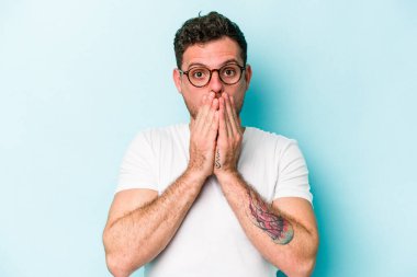 Mavi arka planda izole edilmiş genç beyaz adam elleriyle ağzını kapatırken şok oldu..