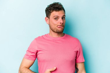 Mavi arka planda izole edilmiş genç beyaz adam karnına dokunuyor, nazikçe gülümsüyor, yemek yiyor ve tatmin oluyor..