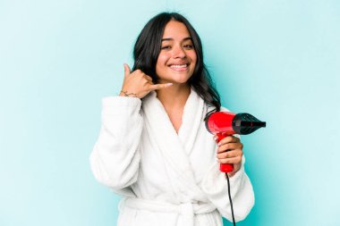 Mavi arka planda izole edilmiş kurutma makinesini tutan genç İspanyol kadın parmaklarıyla bir cep telefonu hareketi gösteriyor..