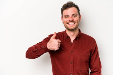 Beyaz arka planda izole edilmiş genç beyaz adam gülümsüyor ve başparmağını kaldırıyor.