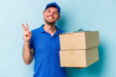 Kafkasyalı genç teslimatçı mavi arka planda izole edilmiş neşeli ve kaygısız parmaklarıyla barış sembolü gösteriyor..