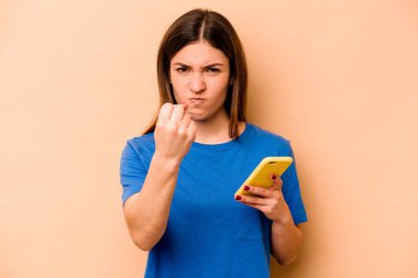Elinde cep telefonu olan beyaz bir kadın bej arka planda izole edilmiş. Kameraya yumruk, agresif yüz ifadesi gösteriyor..