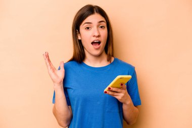Elinde cep telefonu olan beyaz bir kadın bej arka planda izole edilmiş. Şaşırmış ve şok olmuş..