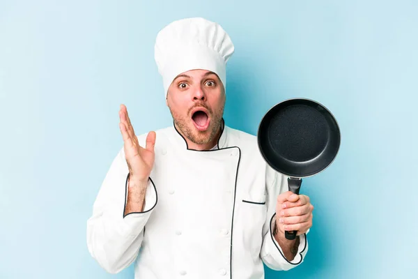 年轻的高加索厨师拿着用蓝色背景隔开的平底锅 惊讶而震惊 — 图库照片