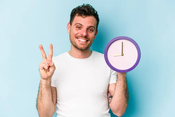 Young Caucasian Man Holding Clock Isolated Blue Background Showing Number — Stock Fotó