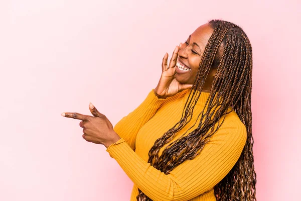 Young african american woman isolated on pink background saying a gossip, pointing to side reporting something.