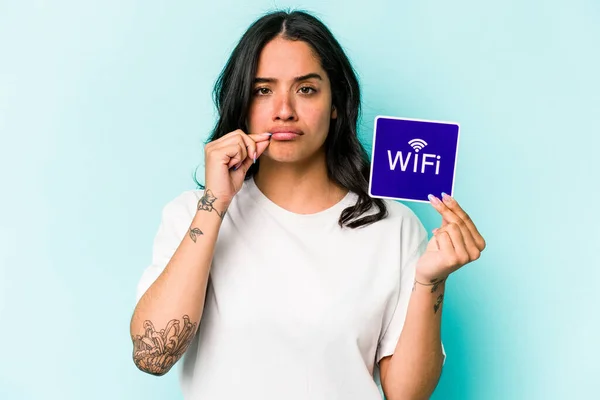 Young hispanic woman holding wifi placard isolated on yellow background with fingers on lips keeping a secret.