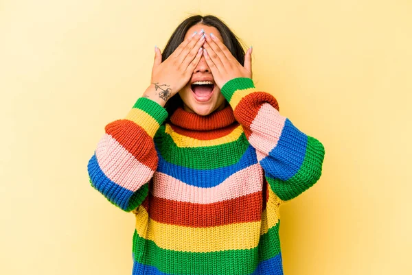 Sarı arka planda izole edilmiş genç İspanyol kadın elleriyle gözlerini kapatıyor, geniş bir gülümsemeyle bir sürpriz bekliyor..