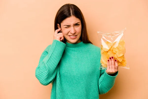 Genç İspanyol kadın elinde bir çanta dolusu cipsle bej arka planda izole edilmiş kulakları elleriyle kapatıyor..