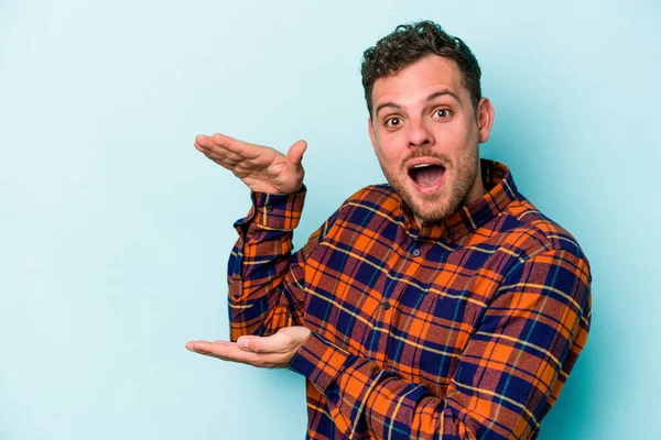 Mavi arka planda izole genç beyaz adam şok ve hayret içinde elleri arasında bir kopya boşluk tutuyor.