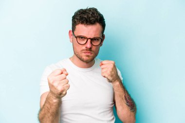 Mavi arka planda izole edilmiş genç beyaz adam yumruğunu kameraya gösteriyor, agresif yüz ifadesi var..
