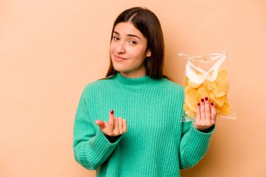 Latin kökenli genç bir kadın, bej arka planda izole edilmiş bir çanta tutuyor. Seni işaret ediyor. Sanki seni davet ediyormuş gibi..
