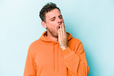 Mavi arka planda izole edilmiş genç beyaz adam esniyor. Ağzı eliyle kapatan yorgun bir hareket sergiliyor..