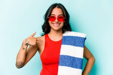 Mavi arka planda izole edilmiş plaj havlusu tutan genç İspanyol kadın gömlek fotokopi alanını işaret ediyor, gururlu ve kendinden emin.
