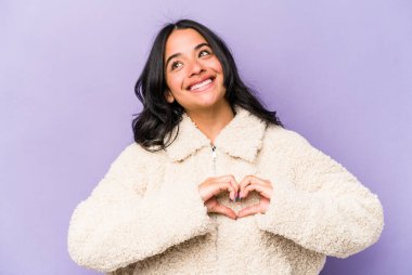 Mor arka planda izole edilmiş genç İspanyol kadın gülümsüyor ve elleriyle kalp şeklini gösteriyor..
