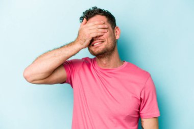 Mavi arka planda soyutlanmış genç beyaz bir adam elleriyle gözlerini kapatıyor, geniş bir gülümsemeyle bir sürpriz bekliyor..