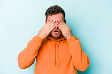 Mavi arka planda izole edilmiş genç beyaz adam elleriyle gözlerini kapatmaktan korkuyor..