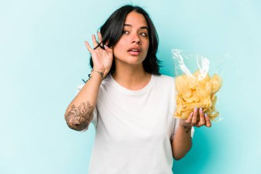 Mavi arka planda bir çanta dolusu cips tutan genç İspanyol kadın bir dedikoduyu dinlemeye çalışıyor..