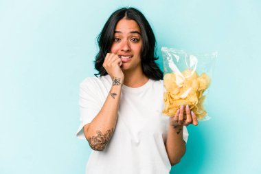 Mavi arka planda izole edilmiş, tırnak yiyen, gergin ve endişeli bir çanta dolusu cips tutan genç İspanyol kadın..