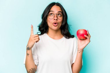 Mavi arka planda izole edilmiş bir elma tutan genç İspanyol kadın ağzı açık bir şekilde üst tarafı işaret ediyor..