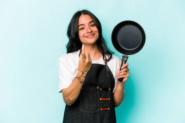 Young hispanic cooker woman holding frying pan isolated on blue background pointing with finger at you as if inviting come closer.