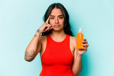 Mavi arka planda güneş kremi tutan genç İspanyol kadın şakağı parmağıyla işaret ediyor, düşünüyor ve bir göreve odaklanıyor..
