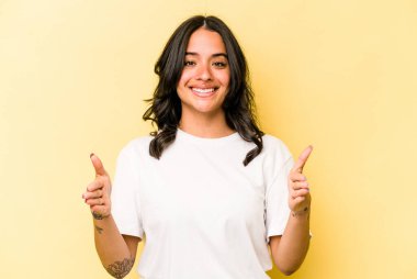 Sarı arka planda izole edilmiş genç İspanyol kadın iki eliyle bir şey tutuyor, ürün sunumu..