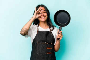 Young hispanic cooker woman holding frying pan isolated on blue background excited keeping ok gesture on eye.