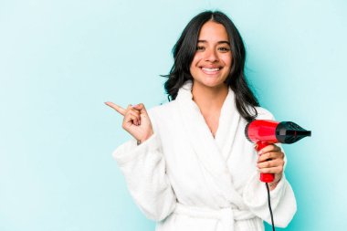 Mavi arka planda izole edilmiş kurutucuyu tutan genç İspanyol kadın gülümsüyor ve boş alanda bir şey gösteriyor..