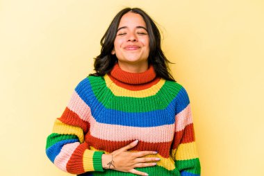 Sarı arka planda izole edilmiş genç İspanyol kadın karnına dokunuyor, nazikçe gülümsüyor, yemek yiyor ve tatmin oluyor..