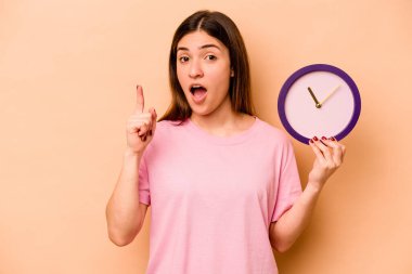 Latin kökenli genç bir kadın bej arka planda izole edilmiş bir saat tutuyor. Bir fikri, ilham verici bir fikri var..
